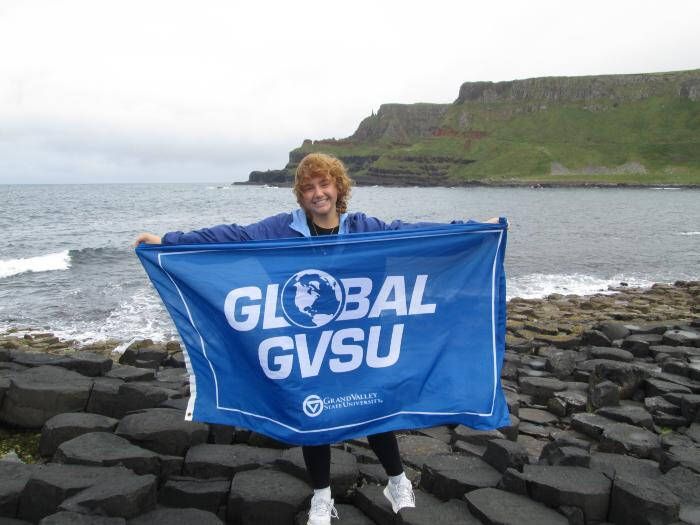 A photograph of a GVSU student holding a Global GVSU flag next to the shore.